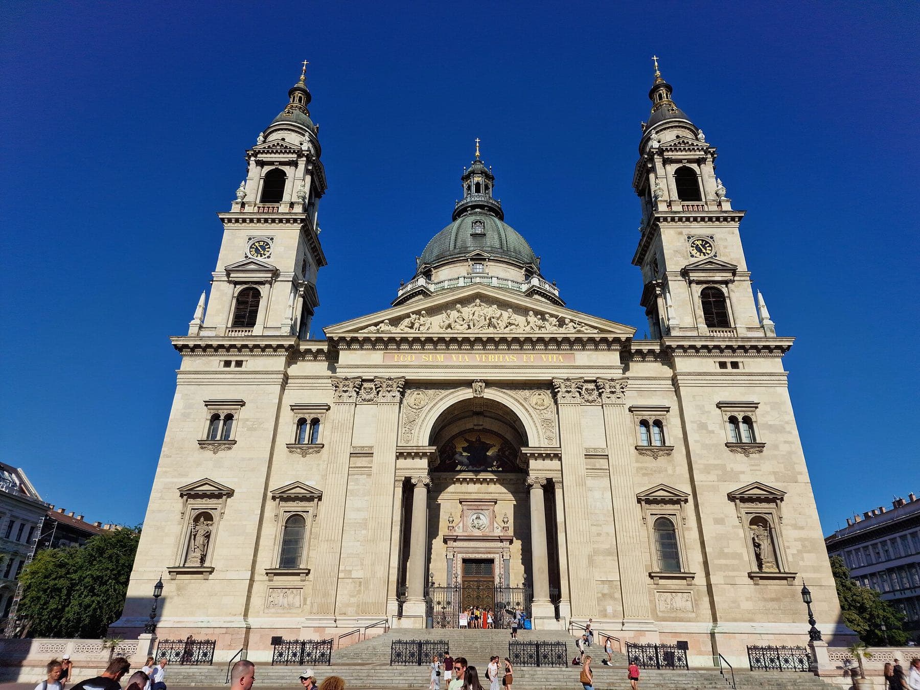 De lichte voorgevel van de Sint-Stefanusbasiliek met twee klokkentorens en de koepel tegen een diepblauwe lucht, met bezoekers op de trappen