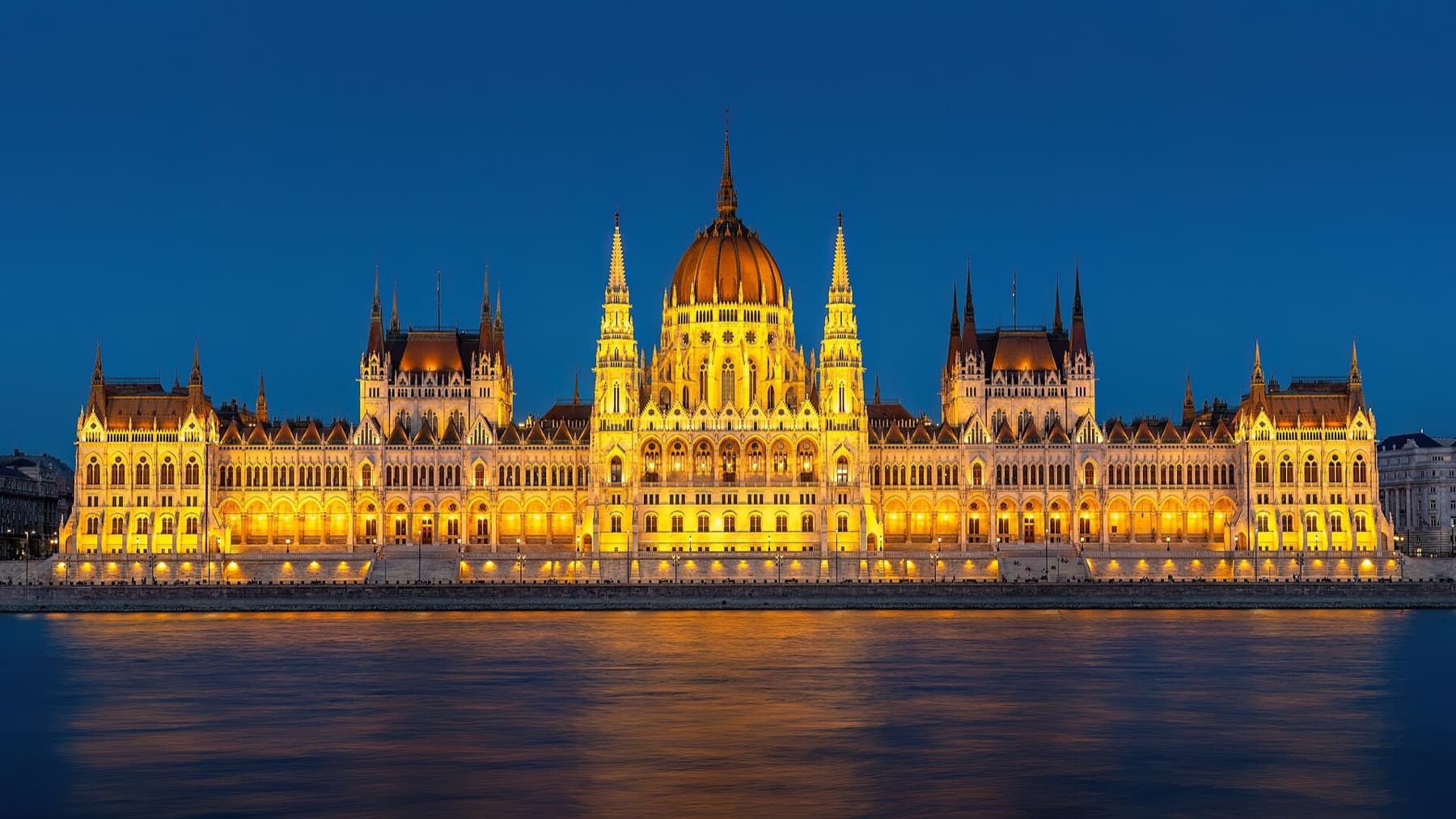 Het verlichte Hongaarse Parlementsgebouw tijdens het blauwe uur, gezien vanaf de overkant van de Donau, met goudkleurige weerspiegelingen in het water