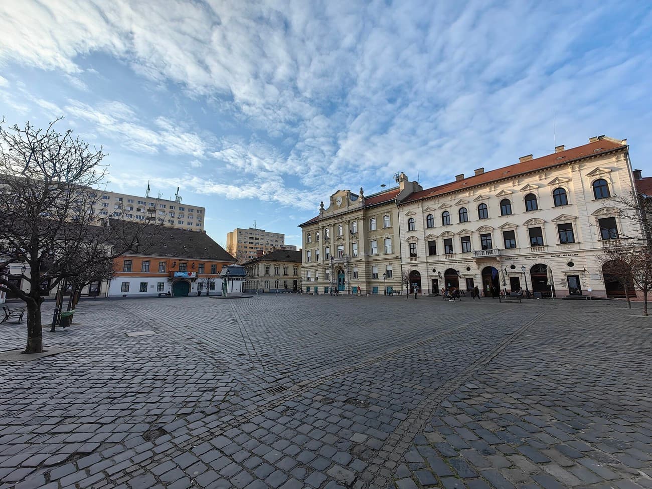 Het ruime, met kinderkopjes geplaveide Fő tér in Óbuda met lichte historische gevels onder een wolkenlucht